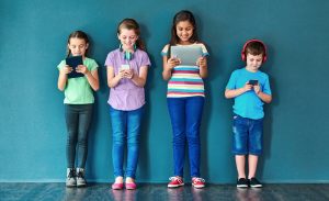 F&uuml;nf Kinder stehen nebeneinander vor einer Wand und nutzen unterschiedliche digitale Endger&auml;te.